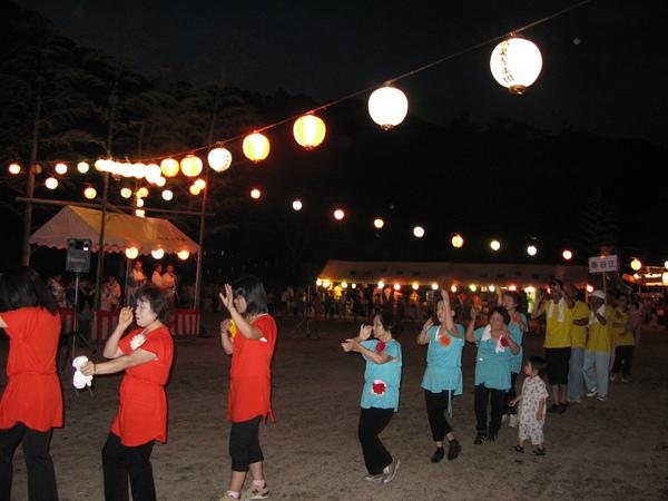 提灯が照らされ、広場では赤・黄・薄ブルーのティシャッを着た人たちが手を上げて踊っている写真