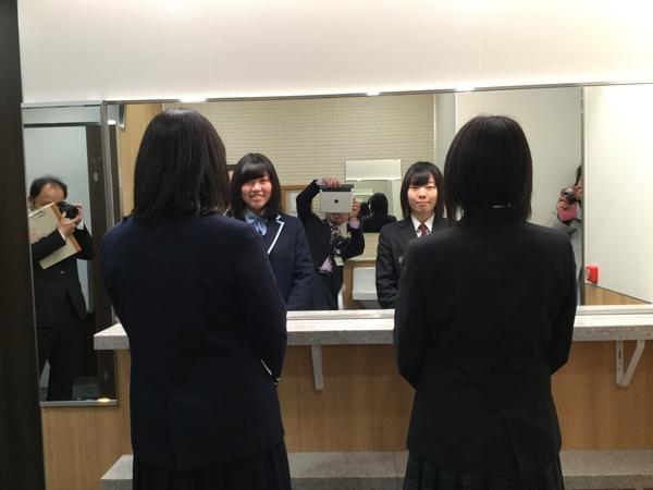 女子高生2人が鏡の前に立ち、周りではスーツを着た男性がタブレットやカメラで写している写真
