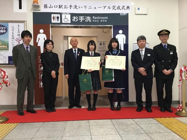 赤いカーペットの上を駅長さん、女子高生2人、市長たちが立って記念撮影写真