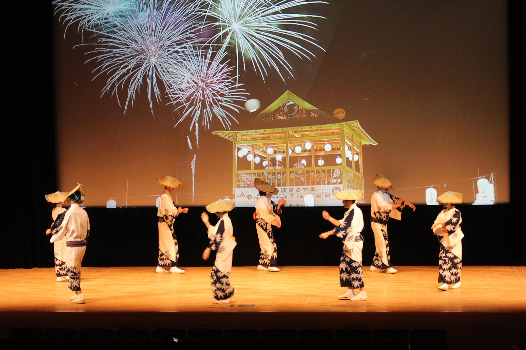 デカンショのやぐらと花火が上がっている景色をスクリーンに映し、7名が舞台で笹浴衣を着てデカンショ踊りを踊っている写真