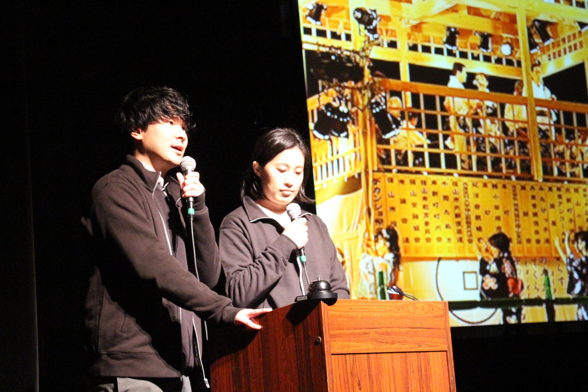 演台のそばでマイクを持って話す市の職員2名の写真