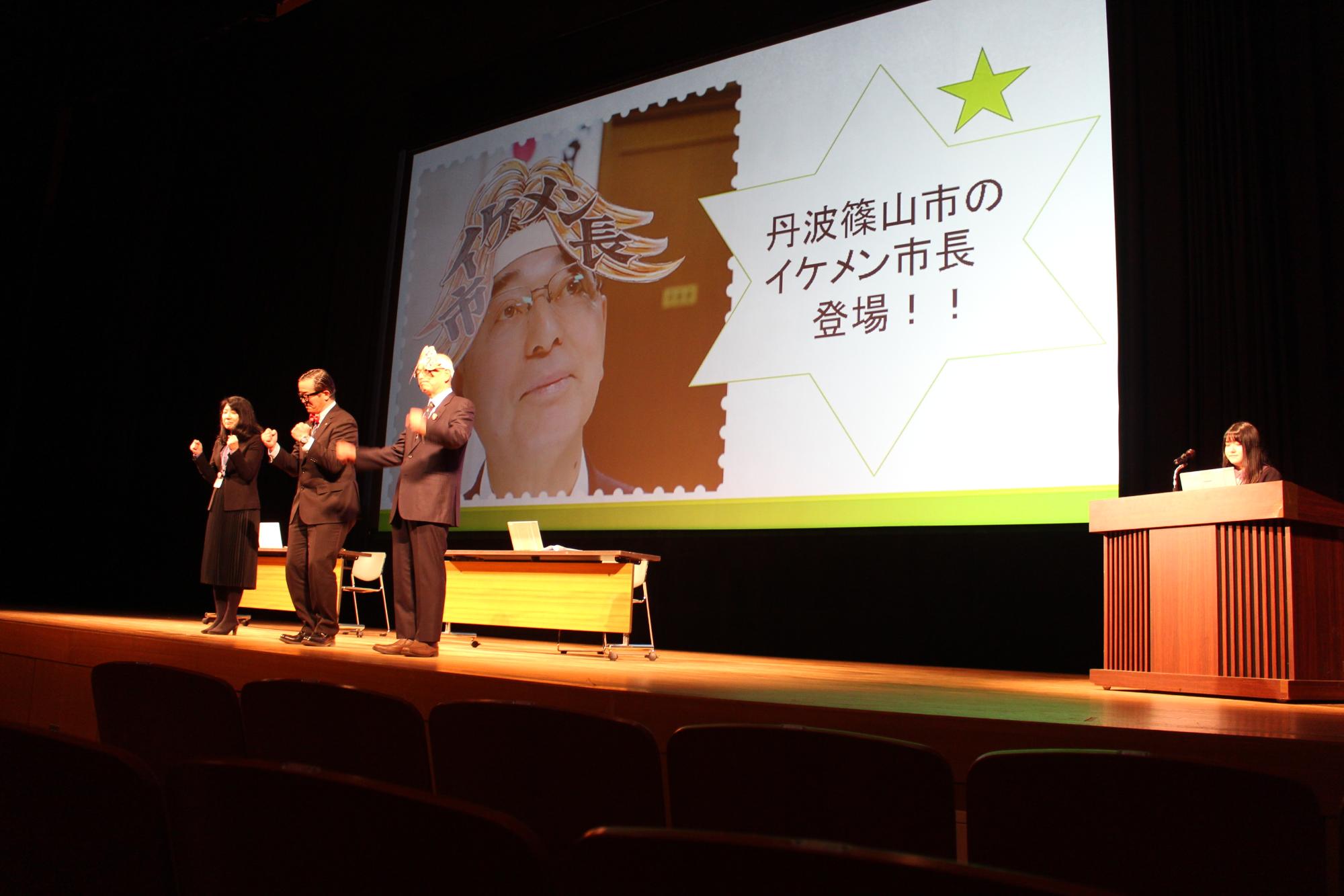 イケメン市長の被り物を被った酒井市長が斜め上を見ている写真をスクリーンに映し、その前で酒井市長と秘書課長と秘書が両手をグーにしてやる気を出している写真