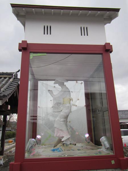 朱色の木枠にガラスがいれられたケースの上に屋根があり中に手をたたいている着物姿のデカンショ人形が入った建設中の写真