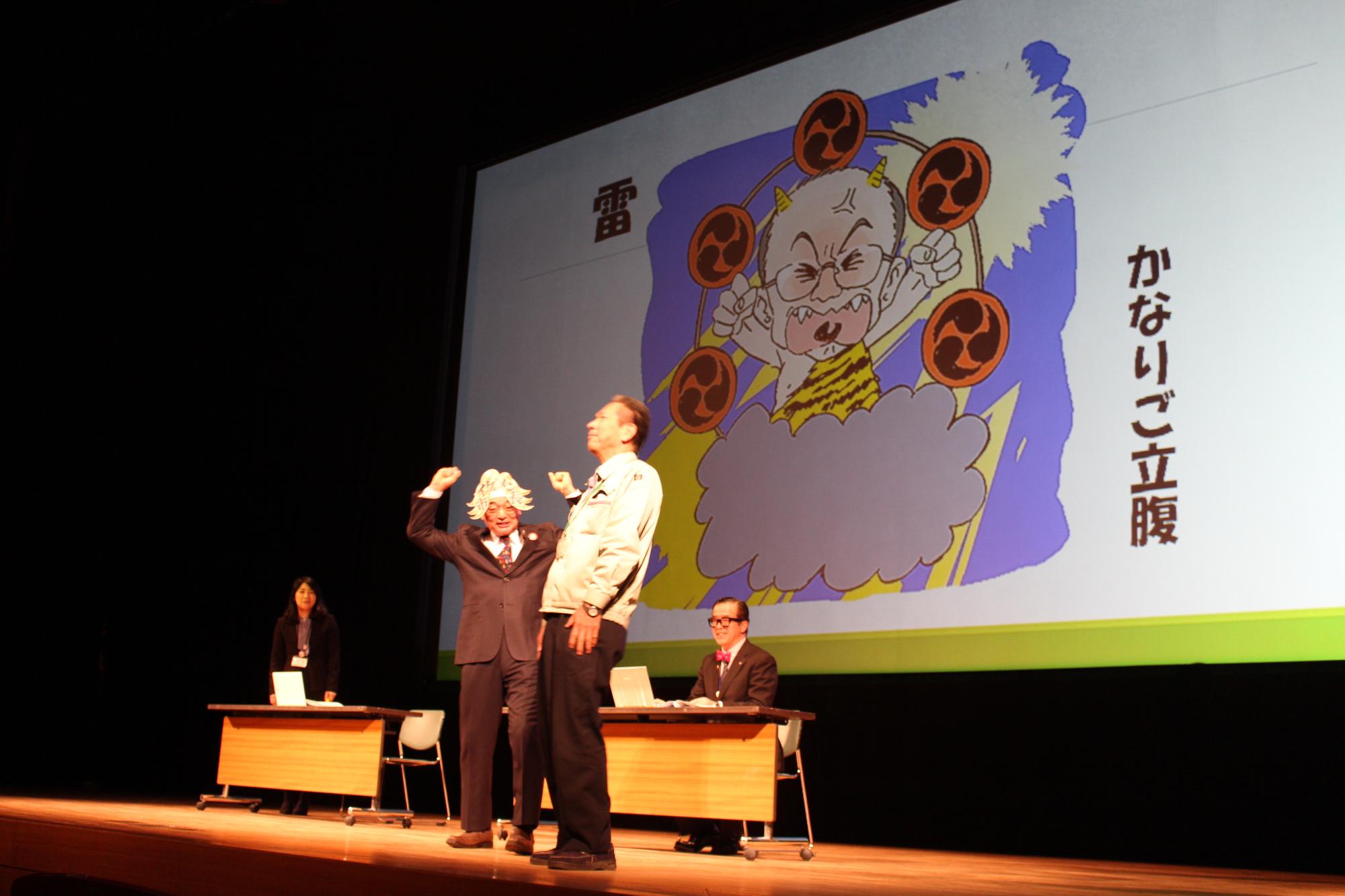 市長のお天気予報の雷マークをスクリーンに映し、職員が立っている横で酒井市長が両手を握って上にあげ、雷さんをしている写真