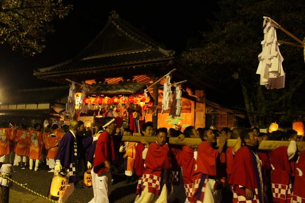 夜、法被姿の人々が神輿を担いで練り歩いている写真