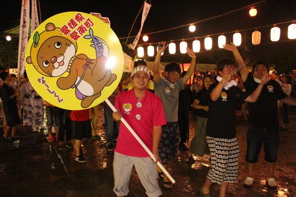 愛媛県愛南町のプラカードを持つ男性と、その後ろで笑顔で踊る男女の写真