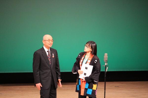 会場の舞台の上で、市長と法被を着た中村秘書がスタンドマイクの前に立って話をしている様子の写真