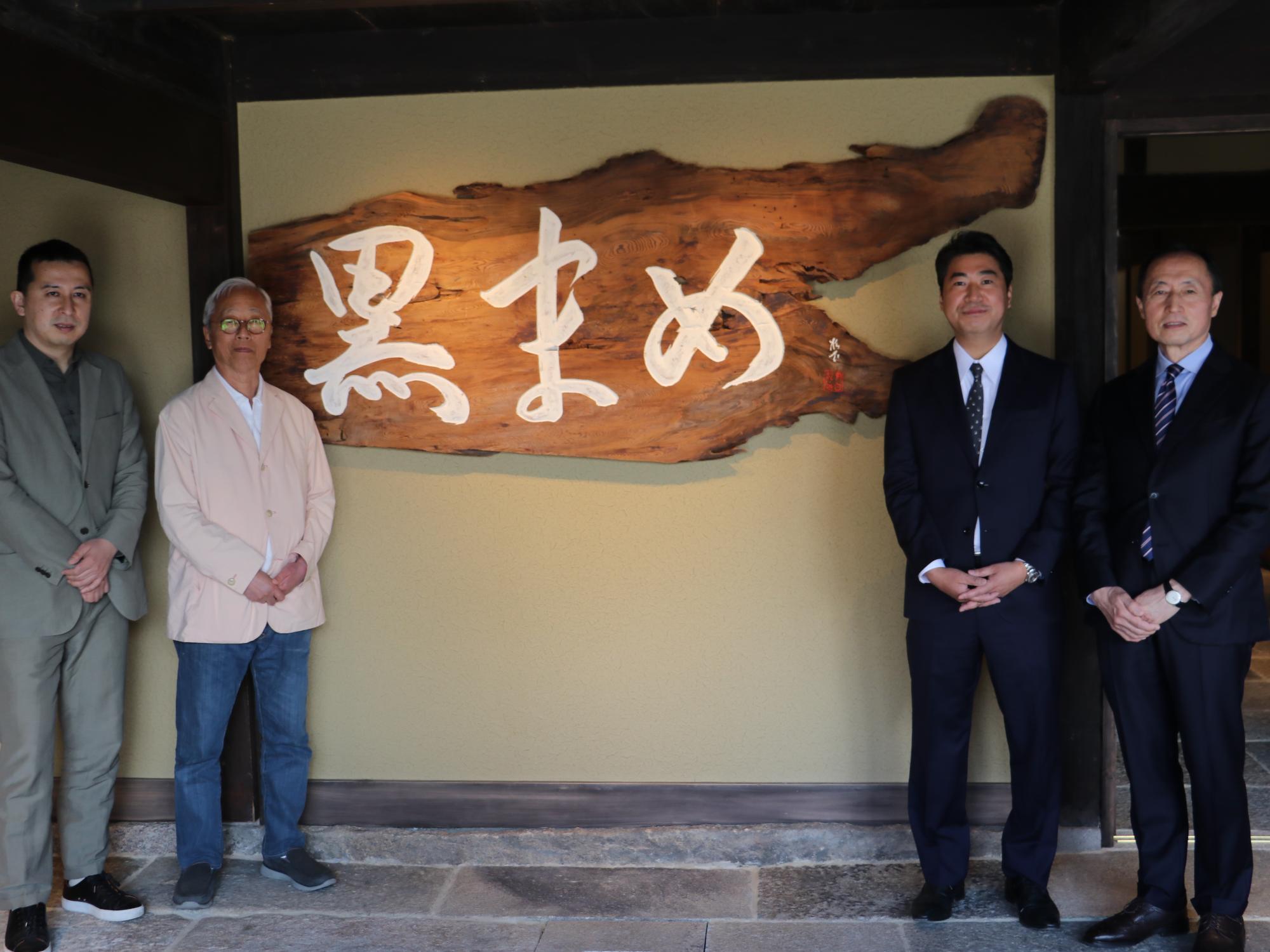 木に白い字で「黒まめ」と書かれた看板のそばに男性4人が立っている。