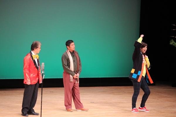 舞台の上で赤いタキシードに金色の蝶ネクタイの男性と白と茶色のマフラーを首にかけた男性が、法被を着た女性が右手を頭の上に上げて踊っているのを見ている様子の写真