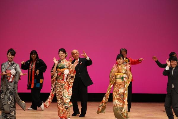 成人式のステージ上で振袖を着た女性3名とスーツを着た男性、後ろに法被を着た実行委員4名が踊っている写真