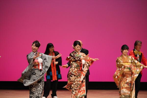 舞台の上で振袖を着た女性3名が両手を前に出して恋ダンスを踊っている様子の写真