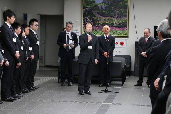 中央の男性がマイクを持って話をしており、市長や整列している職員の方が話を聞いている様子の写真