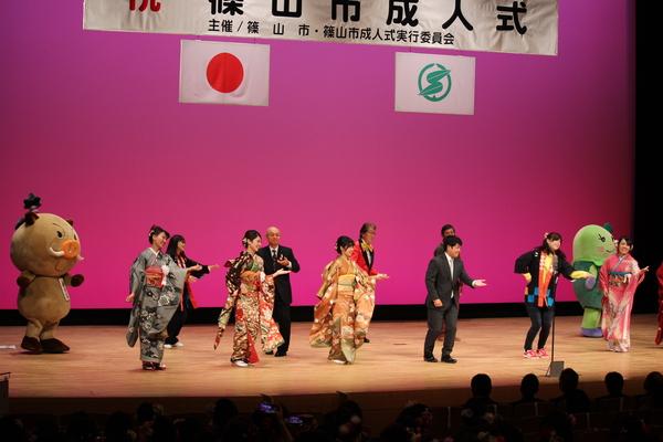 篠山市成人式の横断幕が下がっている舞台上に、振袖を着た女性4名とスーツを着た男性、後には「まるいの」、市長、「まめりん」が一緒にに左手の手のひらを上にして恋ダンスを踊っている様子の写真