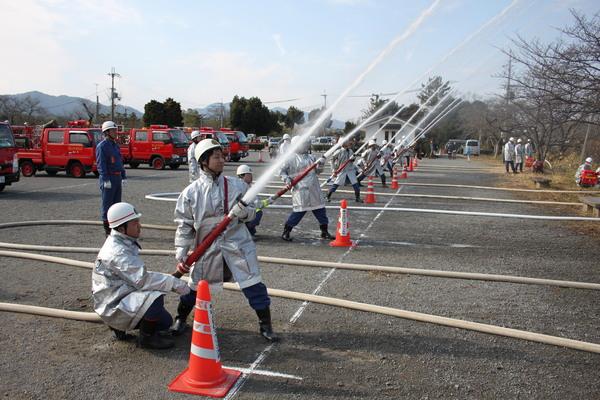 消防団員が二人一組で消防車のホースを持ち、放水演習を行っているのを右側から撮影されている写真