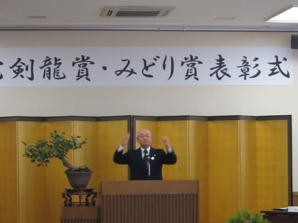 表彰式で金屏風の前にたち市長が両手を上げて挨拶をしている写真