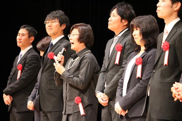 胸に赤い胸章を付けたスーツをきた男性と女性が数名舞台上に並んで立っており、メガネをかけた女性がマイクを持って話をしている写真