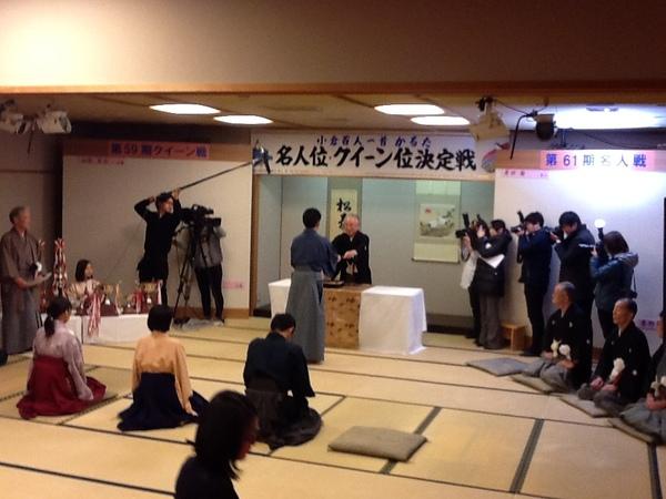 和室でかるた大会の表彰を授与している袴姿の岸田 諭さんをカメラマンが撮影している様子を来賓者と出場者の正座して見ている写真