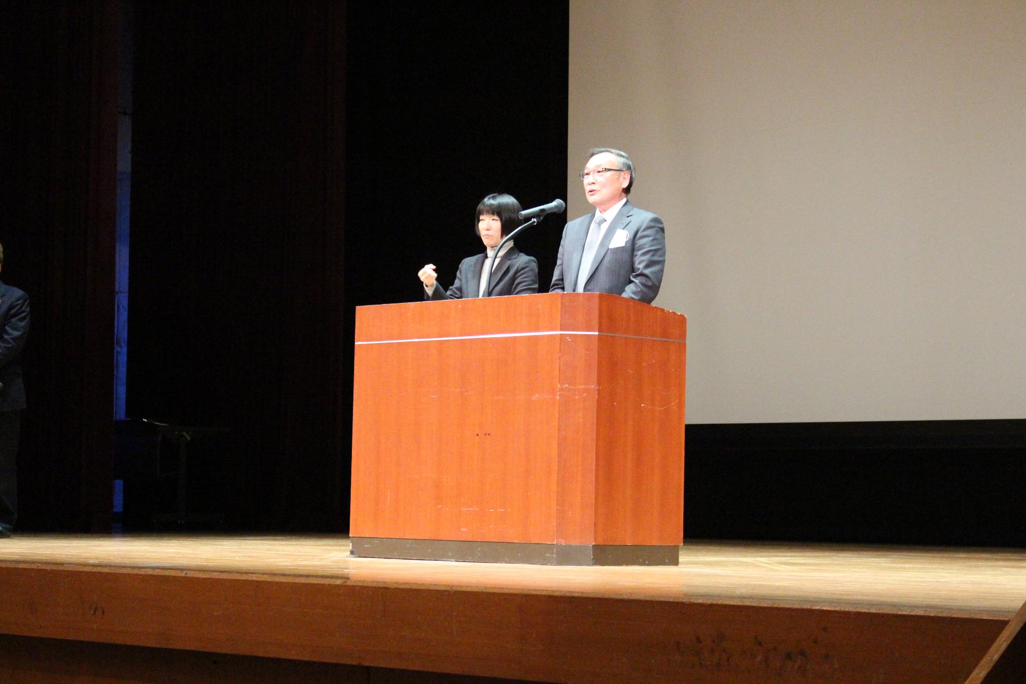 演台の後ろに立って話をする西潟自治会長会会長と、その横で手話をしている女性の写真