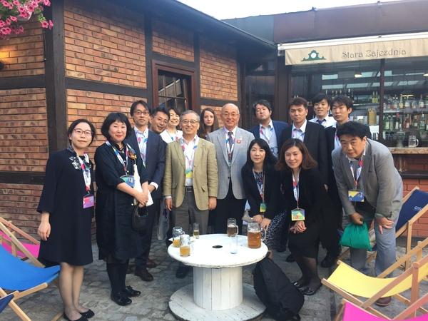 首にネームプレートを下げた15名の男女が丸いテーブルの周りに集合している写真