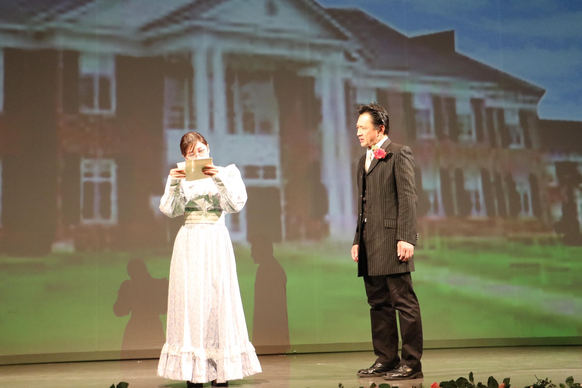 緑の芝生と2階建ての大きな家がスクリーンに映されている前で、白いワンピースのようなものを着た若き日のサリバン先生が何かを読んでいる横で、背広姿の男性が話しかけている写真