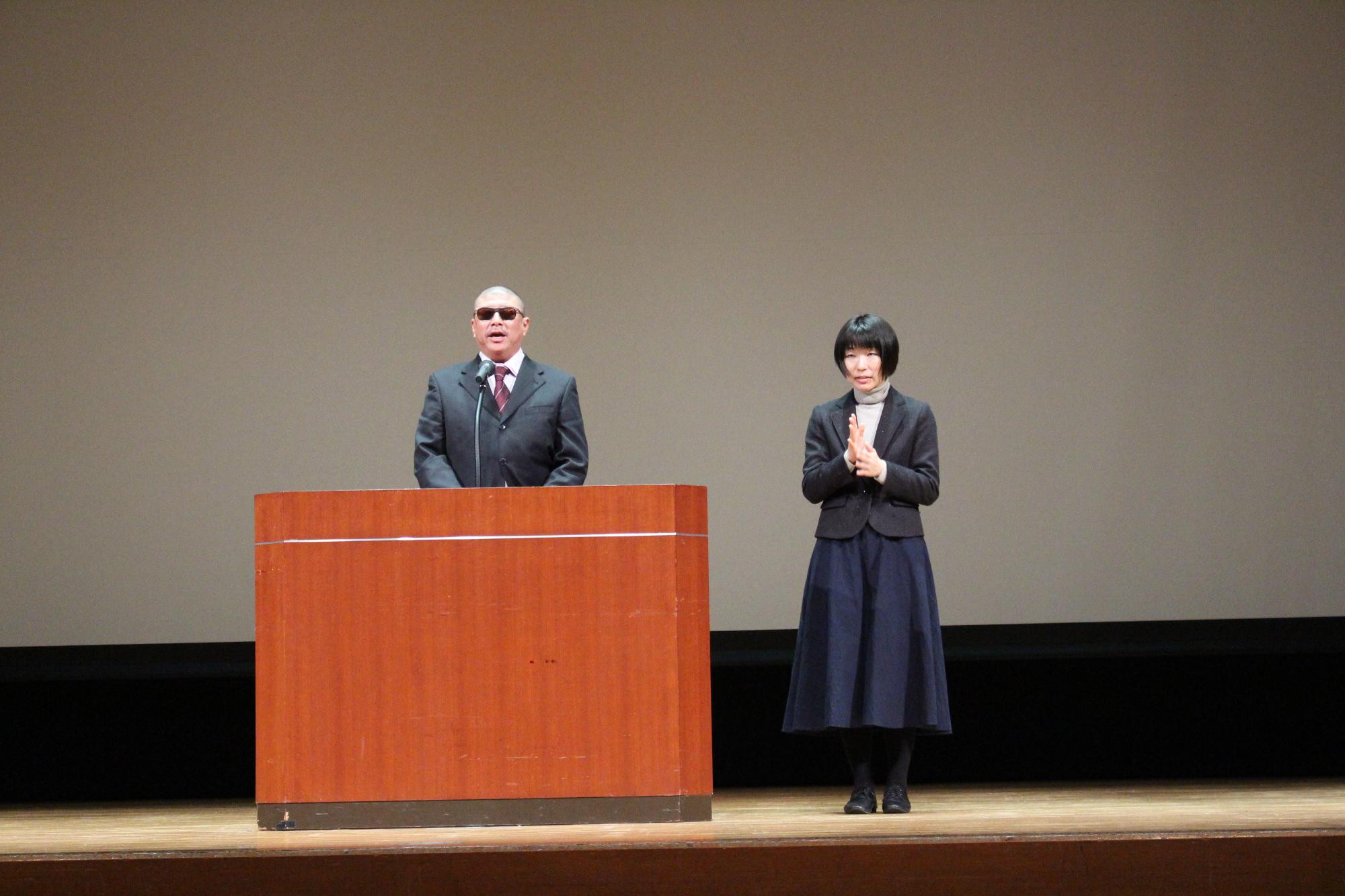 演台の後ろに立って話す辻本会長と、その横で手話をしている女性の写真