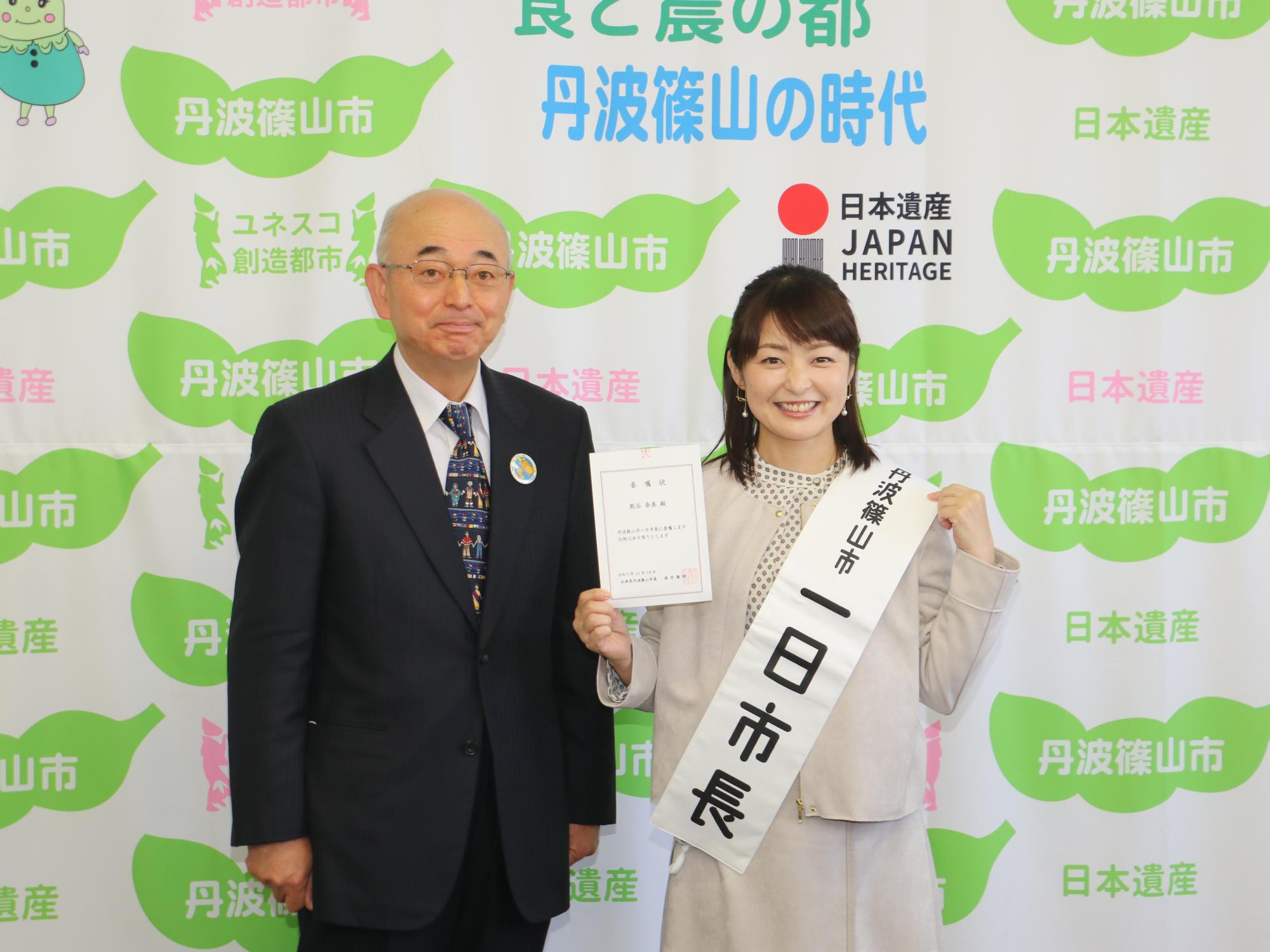 丹波篠山市一日市長というたすきをかけて、右手に委嘱状を持った熊谷市長と、酒井市長の記念撮影