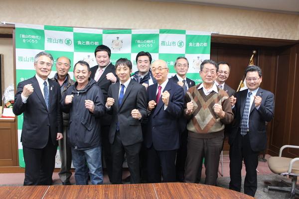 前列でガッツポーズをしている角谷選手と市長が笑顔で写っており、その周りに関係者の方々も一緒にガッツポーズをして写っている集合写真