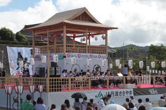 祭りの舞台の上で吹奏楽部の生徒さんがお客さんの前で演奏している写真