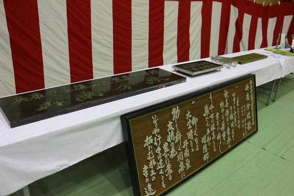 紅白の幕が張られ、長机には白い布が被せられ、その上に文字が書かれた石碑が置いてある写真