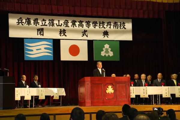 ステージ上の演台前に立ち、話をしている大前さんとその後方には関係者の方々が椅子に座っている写真