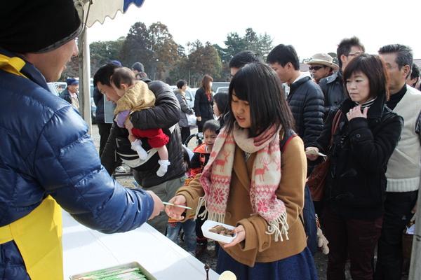 エプロン姿の男性がマフラーを巻いた女の子に料理の入った紙皿と割りばしを渡している写真