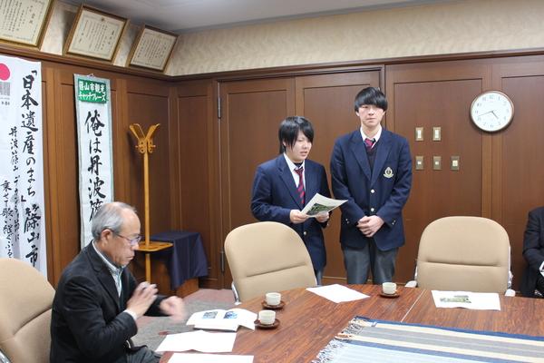 紺色のブレザーを着ている高校生2名が立っており、左側の生徒が原稿を手に持って読んでおり、椅子に座ってメガネをかけた男性が机の上に置かれた資料を興味深く見ている様子の写真