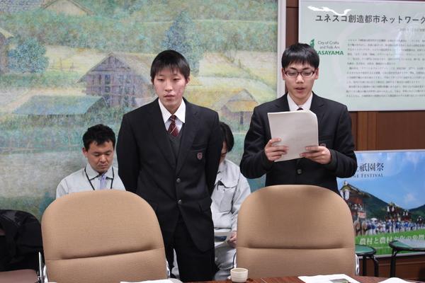 黒いブレザーを着ている高校生2名が立っており、右側のメガネをかけている生徒が手にもった原稿を読んでいる様子の写真