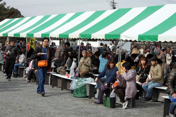 緑と白のボーダーのテントの下に沢山の人達が座っていて、食べ物を食べたり、話をしている写真