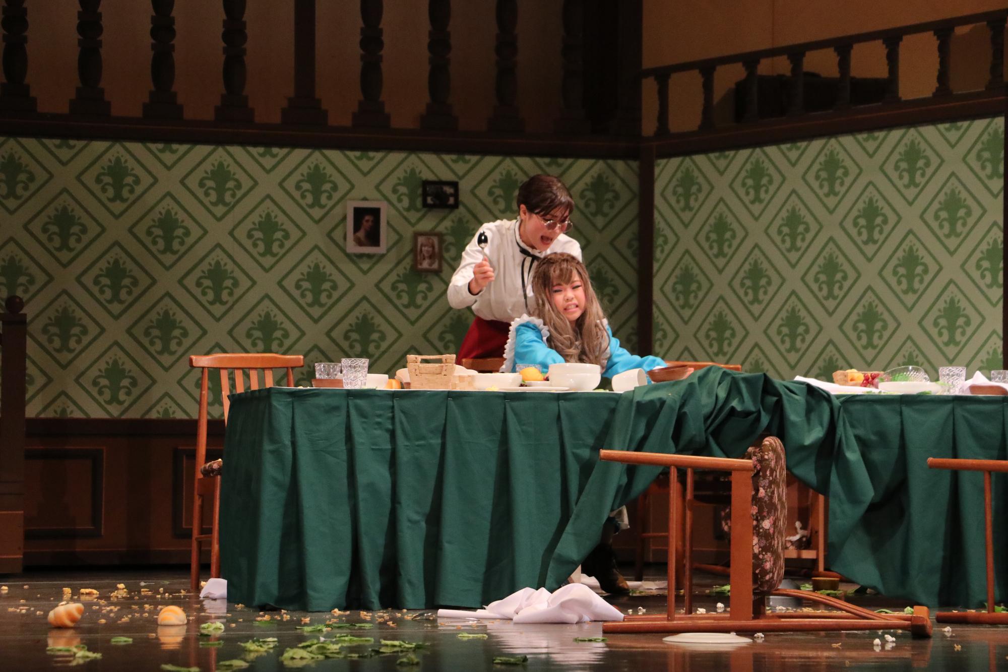 緑のテーブルクロスがひいてある机に食事が用意されていたが、イスは倒され、パンなどの食事はひっくり返って床に落ちている。座っている幼いヘレンの後ろで厳しく注意するサリバン先生