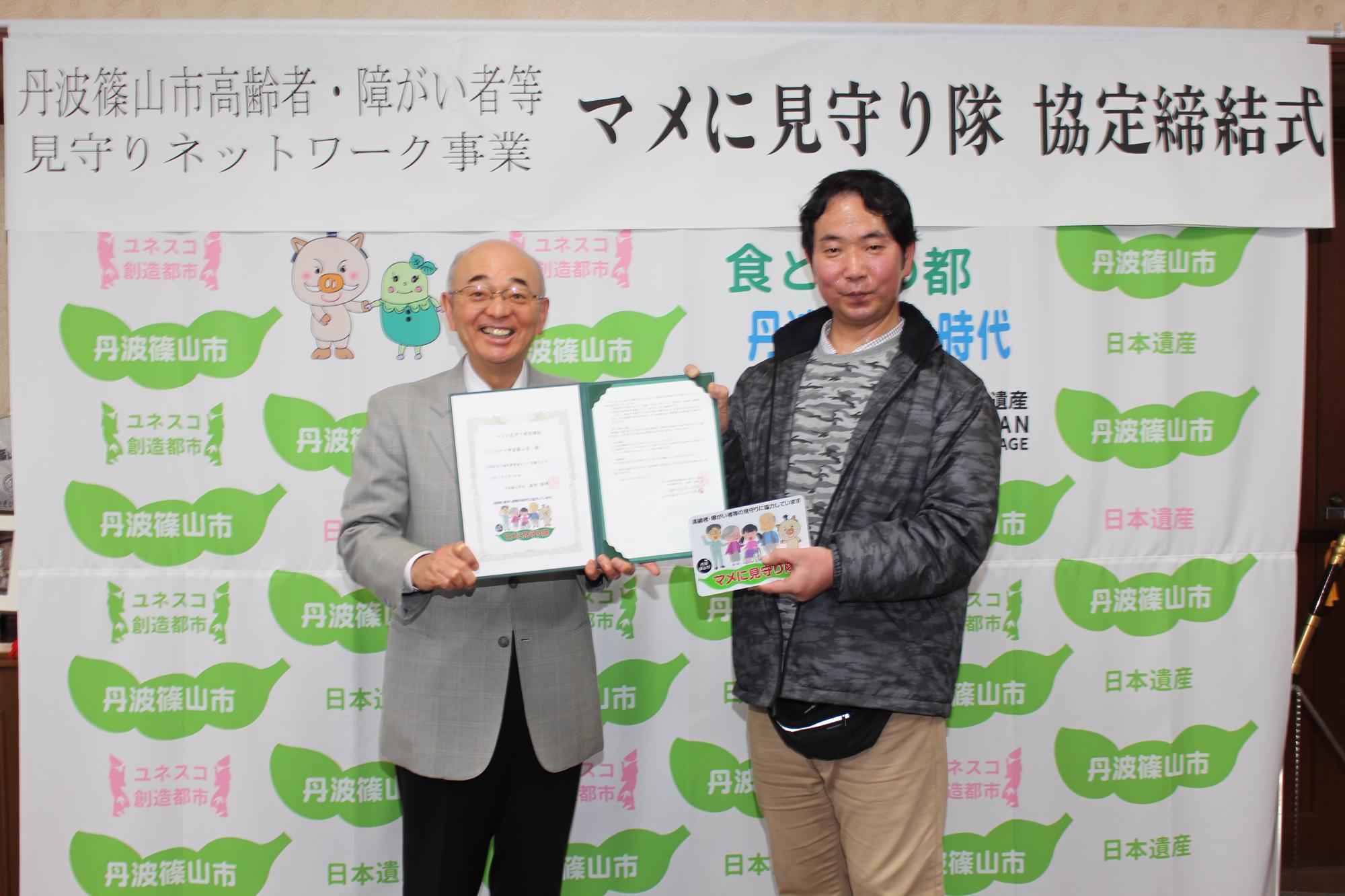 マメに見守り隊締結式で、酒井市長と杉本さんがマメに見守り隊のステッカーなどを持って横に並んでいる記念写真