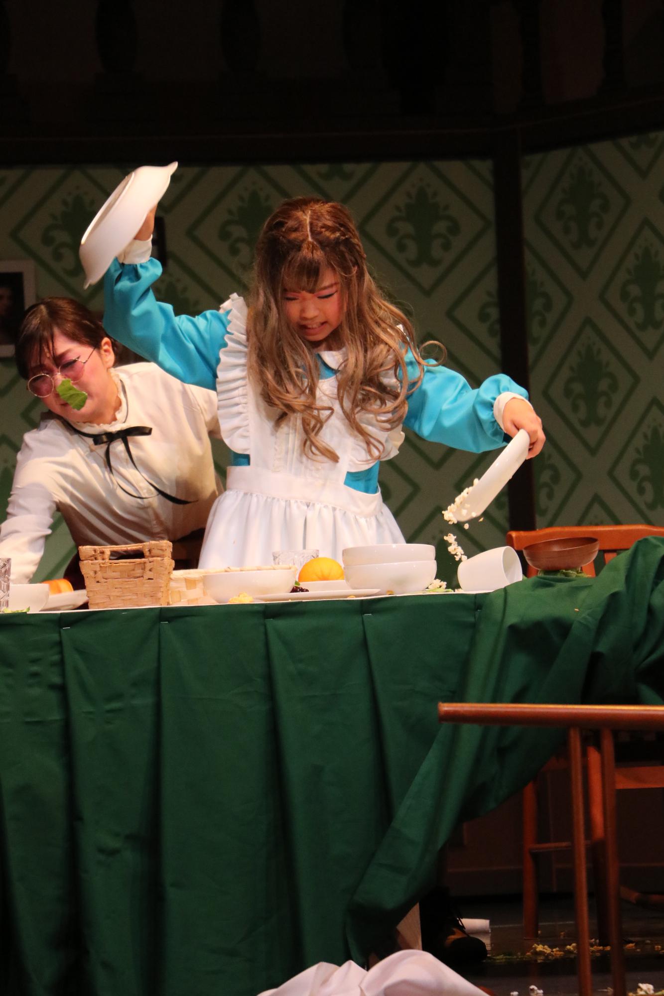 水色のブラウスに白のエプロンを付けたヘレンが、食べ物が入ったお皿などをひっくり返している後ろで、それを止めようとするサリバン先生の写真