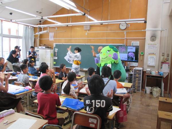 黒板の前で市長や浴衣を着た女性、着ぐるみが踊っているのを生徒達が拍手をしながら見ている様子を窓際から写真を撮っている人がいる教室内の写真