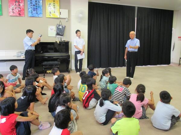 床に子供たちが座っていて、立ちながらビデオで撮影している人、直利不動でたっている人の横で市長が立って話をしている様子の写真