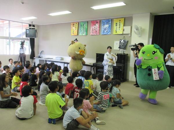 教室の床に座った子ども達の周りをまめりん、まるいのの着ぐるみ、浴衣を着た女性が踊っている様子を全体的に写した写真