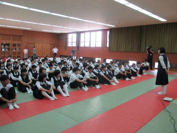 赤と緑の交互の床に50名ほどの中学生が座っていて、代表の女子中学生が前に立って話をしている写真