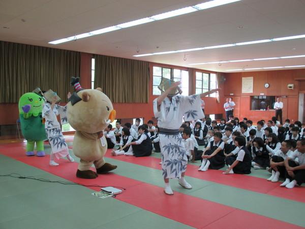 前列で浴衣を着た女性と男性とまるいの、まめりんの着ぐるみが踊っている姿を制服姿の生徒達が座って笑顔でみている写真