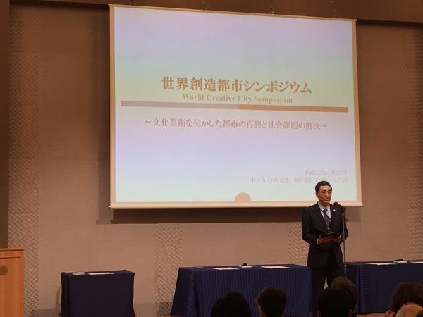 文化庁の磯谷 桂介氏が、スタンドマイクを使い聴衆のまえで挨拶している写真