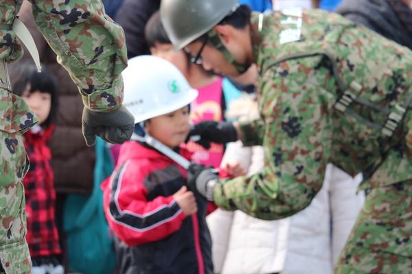 自衛隊員の男性が、男の子の白いヘルメットの首の留め具を付けている様子の写真