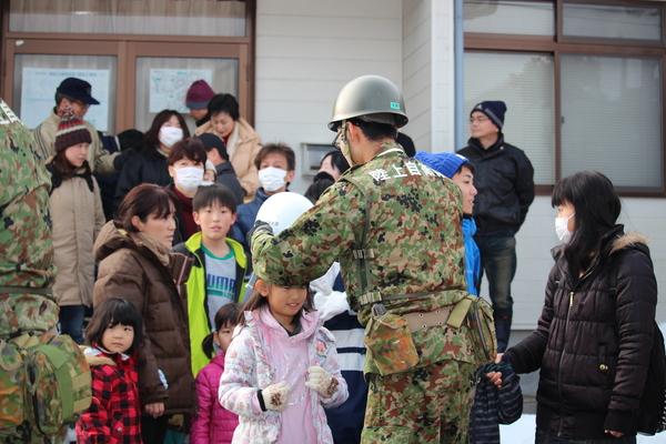 陸上自衛隊員の男性の前に親子連れの住民が並んでおり、ピンクの洋服を着ている先頭の女の子にヘルメットをかぶせてあげている様子の写真