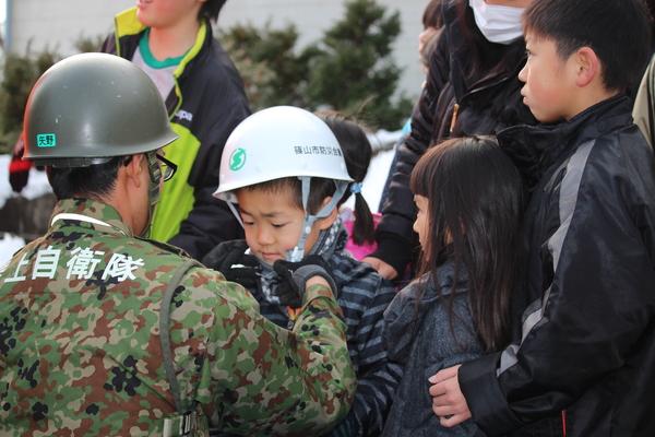 陸上自衛隊員の男性の周りに親子連れが集まっており、小さい男の子に膝を下ろしてヘルメット付けてあげている様子の写真