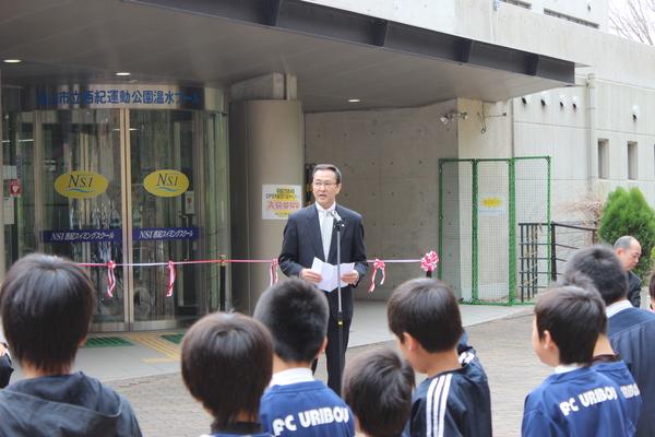 近藤 雅彦社長スタンドマイクの前で話をしており、後にはテープカットの紅白のリボンが設置され、男子児童達が話を聞いている後ろ姿が写っている様子の写真