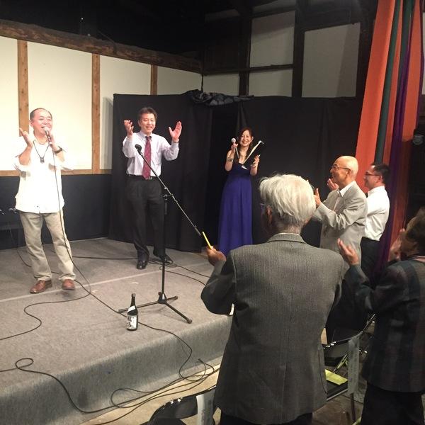 マイクを持った男性が歌を披露、その周りでみんでが手拍子している写真