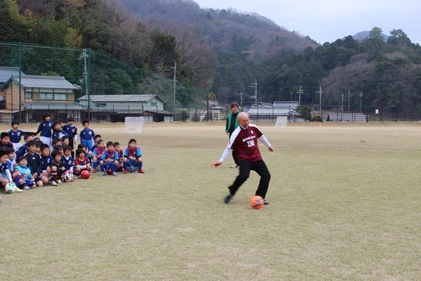 グラウンドにサッカー少年が集まって座っており、市長が子ども達の前でオレンジ色のサッカーボールをけっている様子の写真