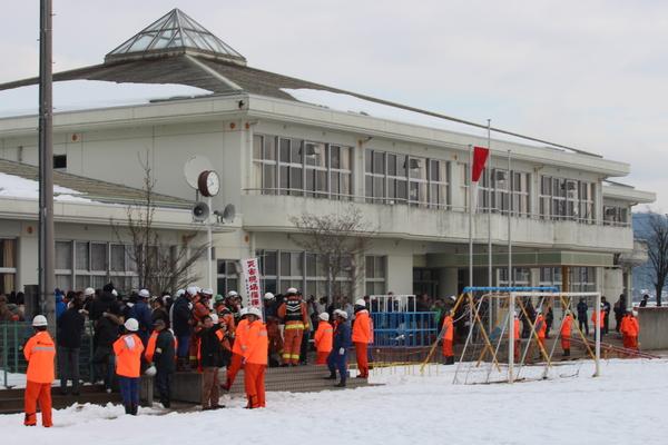 雪の降り積もる中、西紀南小学校の校舎の前に沢山の住民が集まっており、オレンジ色のジャンパーを着た関係者が集まった住民の前に立っている様子の写真